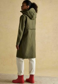 Caban impermeabil de culoare verde măslin, cu glugă, de lungime până la genunchi, cu ventilație pe spate, asortat cu pantaloni albi răsuciți și cizme roșii din cauciuc.