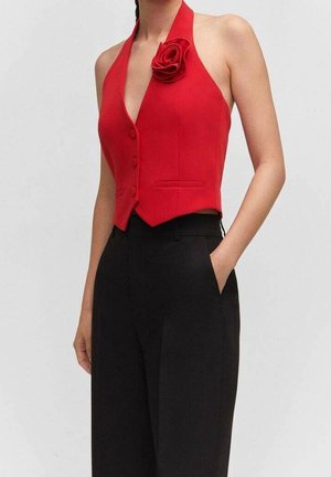 Femme portant un gilet rouge sans manches à col halter avec un détail de rose, associé à un pantalon noir taille haute, les mains dans les poches.