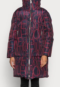 Lång marinblå puffjacka med röd geometrisk bokstavsmönster, framtida dragkedja, dragkedjefickor på sidan och hög krage som bärs av en person.