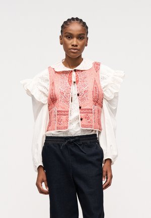 Frau mit geflochtenem Haar trägt eine weiße Bluse mit Rüschen und eine rote Weste mit Muster, kombiniert mit schwarzen Hosen mit Kordelzug vor weißem Hintergrund.