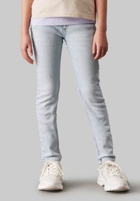 Világoskék farmer skinny nadrág, klasszikus öt zsebes dizájnnal és enyhe kopással. Fehér sportcipővel párosítva.