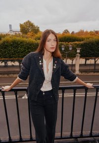 Grauer Wollcardigan mit floraler Stickerei am Kragen, kombiniert mit einer weißen Bluse und schwarzen Hosen, vor einem verschwommenen städtischen Hintergrund.