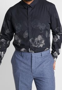 Homme portant une chemise noire boutonnée avec des motifs floraux gris foncé et un pantalon texturé bleu clair, mains dans les poches.