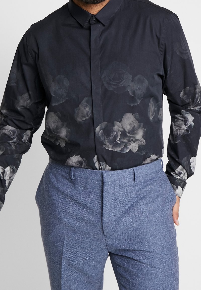 Homme portant une chemise noire boutonnée avec des motifs floraux gris foncé et un pantalon texturé bleu clair, mains dans les poches.