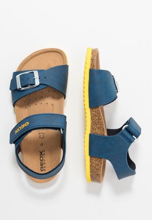 Paire de sandales bleues avec sangles réglables, semelles en liège et semelles en caoutchouc jaune, vues de dessus et de côté sur fond blanc.