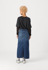 T-shirt noir superposé à un haut transparent à manches longues, associé à une jupe maxi en denim bleue avec une fente arrière et des poches. Baskets blanches aux pieds.