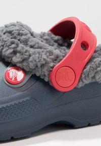 Granatowy chodak z szarym, puszystym wykończeniem, czerwonym paskiem i guzikowymi akcentami. Teksturowana powierzchnia z okrągłymi perforacjami w pobliżu pięty.