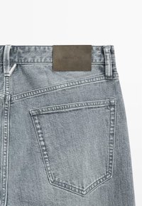 Jean en denim gris avec une texture lisse, doté d'une étiquette carrée en cuir marron marquée "Massimo Dutti" sur la ceinture arrière et de poches standards.