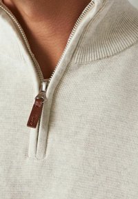 Half-zip sweater in soft beige knit. Features a ribbed collar and a metal zipper with a brown leather pull tab. Simple, textured fabric.