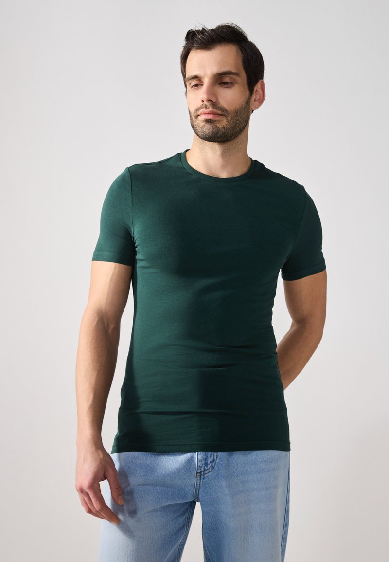 Zielony t-shirt z krótkim rękawem i okrągłym dekoltem, wykonany z gładkiego materiału, dopasowany krój, noszony z jasnoniebieskimi dżinsami. Brak widocznych wzorów.