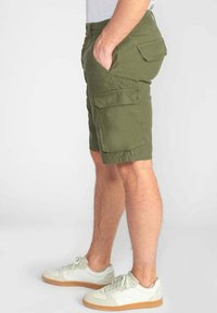 Shorts cargo vert olive avec plusieurs poches à l'avant et sur les côtés, fabriqués en tissu léger. Associés à des baskets blanches.