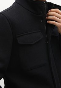 Musta vetoketjupusero, joka on valmistettu teksturoidusta kankaasta. Tässä puserossa on kaksi rintataskua läpällä ja korkea kaulus, jota korostavat painonapit.