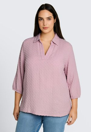 Jeune femme aux longs cheveux foncés portant un chemisier texturé violet clair à manches 3/4 et un jean bleu clair, debout devant un fond uni.