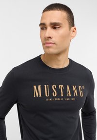 Zwarte lange mouwen t-shirt met een gouden logoafdruk dat "MUSTANG JEANS COMPANY SINCE 1932" leest, zachte katoenstructuur.