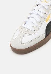 Vita sneakers med grå mockadetaljer och brun gummisula; har platta vita snören och en subtil logotyp i gult på sidan.