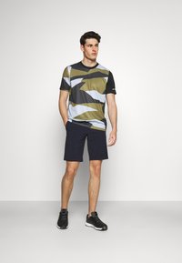 Camisa atlética de manga corta en color verde oliva, con un patrón geométrico en gris y negro. Combinada con pantalones cortos en azul marino. Modelo lleva zapatillas negras.
