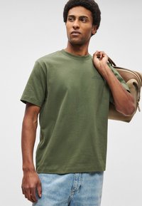 T-shirt de algodão verde-oliva com mangas curtas, decote redondo e um pequeno logótipo azul no peito. Usada com calções de ganga azul claro.