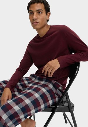 Camicia bordeaux a maniche lunghe abbinata a pantaloni da pigiama a quadri in rosso, bianco e blu navy, seduto su una sedia nera. Tessuto morbido, vestibilità comoda.