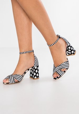 Sandalias de tacón alto en blanco y negro con patrones de rayas y lunares, diseño de tiras cruzadas y correa en el tobillo.