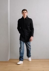 Lungo cappotto nero con colletto, chiusura con bottoni, abbinato a jeans azzurri e sneakers bianche. Texture liscia, design semplice e colori neutri.