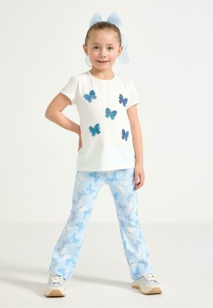 Pige smiler med hænderne på hofterne, iført hvid skjorte med blå sommerfugleapplikationer, lyseblå bukser med sommerfuglemønster og sneakers.