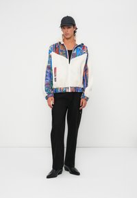 Fleece zip jacket with colorful abstract patterns, cream body, and blue sleeves. Paired with black cargo pants and black shoes.