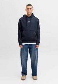 Laivastonsininen huppari kengurutaskulla ja pienellä logolla rinnassa, yhdistettynä väljään siniseen farkkuun ja vaaleanbeigeihin kenkiin.