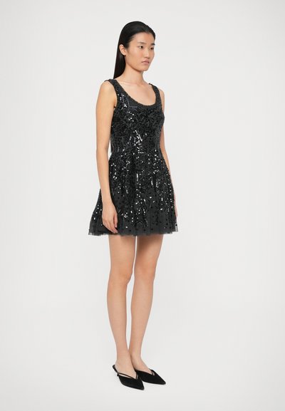 Žena stojící v bezrukávových černých flitrových mini šatech a černých špičatých sandálech na podpatku, na jednolitém světlém pozadí.