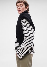 Camisa de mangas longas com riscas em preto e branco, combinada com um suéter de malha preto colocado sobre os ombros; corte casual com costuras visíveis.