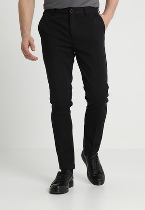 Uomo che indossa pantaloni neri slim fit e scarpe nere stringate, con una mano in tasca, cammina su uno sfondo bianco semplice.