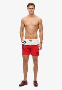 Shorts de bain rouges avec une ceinture blanche, ornés de accents noirs et des chiffres "03" et "23". Fabriqués en matériau léger, adaptés aux activités aquatiques.