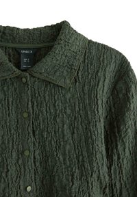 Grøn, struktureret skjorte med krave, frontknapper og et krøllet stofdesign. Mærket læser "Lindex," lavet i Tyrkiet.