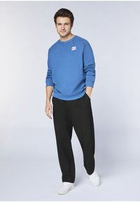 Blauer Sweatshirt mit gerippten Bündchen und Saum, mit einem kleinen Etikett auf der linken Brust. Schwarze, weit geschnittene Hose und weiße Turnschuhe.