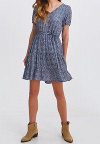 Kobieta ubrana w niebieską sukienkę z krótkim rękawem w wzory, z elastycznym paskiem w talii, w połączeniu z beżowymi zamszowymi botkami do kostek, stojąca na tle jednolitym.