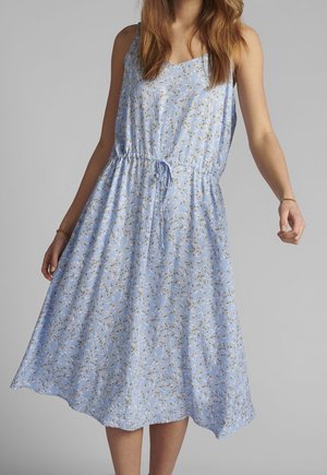 Woman wearing sleeveless light blue midi dress with pastel leaf and dotted pattern, cinched waist with drawstring, standing on gray background.