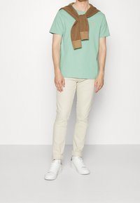 Svetle zelené bavlněné tričko s malým logem, zkombinované s krémovými kalhotami a bílými teniskami. Hnědý pletený svetr je přehozený přes ramena.