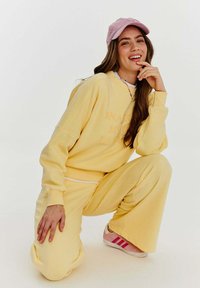 Žlutá mikina se textem "SNAKE MO", doplněná o odpovídající kalhoty. Celý outfit dotváří růžová čepice a tenisky s růžovými pruhy. Měkký materiál a volný střih.