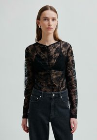 Kobieta w czarnej koronkowej bluzce z długim rękawem na czarnym podkoszulku, w komplecie z ciemnymi dżinsami o podwyższonym stanie, na neutralnym tle.