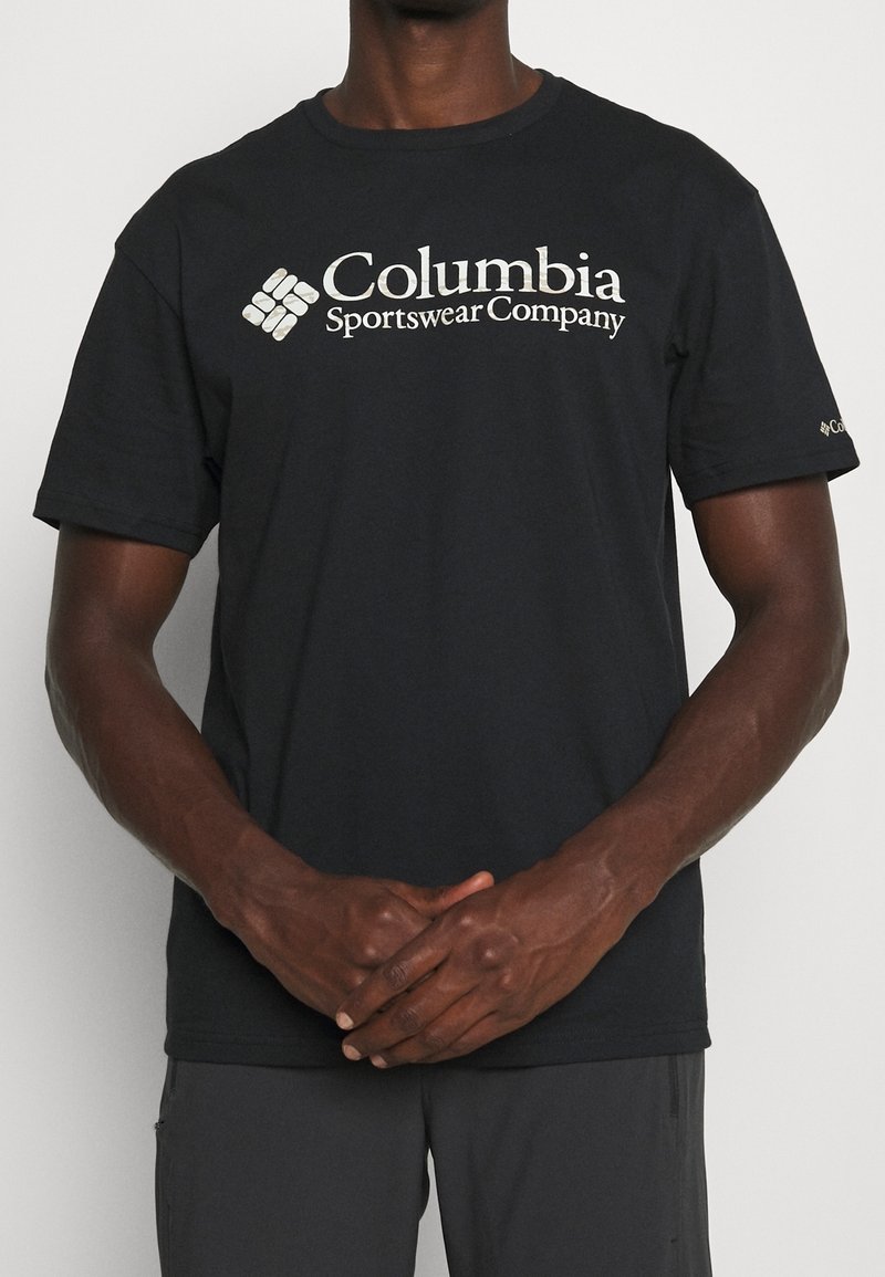 Personne portant un t-shirt noir Columbia Sportswear Company, debout, les mains jointes devant un fond uni.