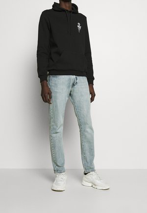 Svart huvtröja med framficka och vit grafisk detalj. Ljusblå jeans med rak skärning. Vita sneakers kompletterar outfiten.