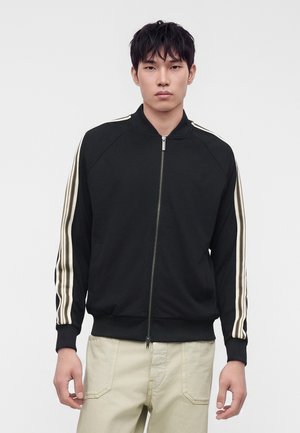 Jeune homme portant une veste noire zippée avec des manches beige rayées et un pantalon beige, debout devant un fond clair uni.