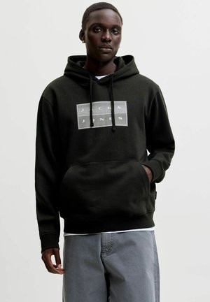 Giovane uomo che indossa una felpa nera Jack & Jones con tasca e pantaloni grigi, in piedi davanti a uno sfondo semplice.