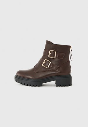 Bottine en cuir marron avec boucles dorées, fermeture zippée et semelle en caoutchouc noir robuste. Présente une finition texturée et une silhouette épurée.