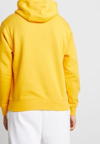 Suéter amarillo con capucha, con una textura suave y una cinturilla acanalada. El diseño de la parte trasera es simple, sin logotipos ni patrones visibles.