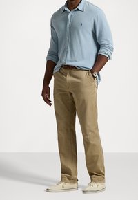 Ljusblå långärmad skjorta med knappar och en diskret logotyp, kombinerad med ljusbeige byxor och beige skor. Har ett vävt bälte och klocka.