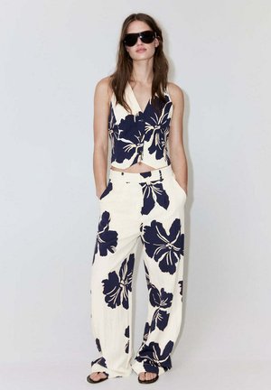Femme portant un gilet sans manches crème et un pantalon à jambes larges avec un grand imprimé floral bleu foncé, des lunettes de soleil noires et des sandales noires, debout les mains dans les poches.