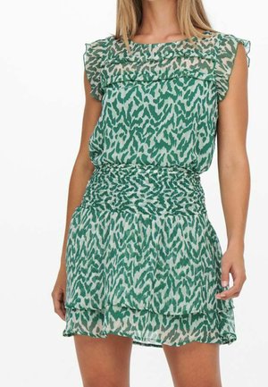 Femme portant une robe sans manches à motifs verts et blancs avec des épaules volantées et une taille froncée, debout devant un fond blanc.