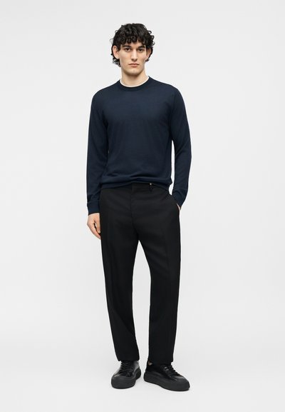 Homme aux cheveux bouclés portant une chemise à manches longues bleu marine, un pantalon noir et des baskets noires, debout devant un fond clair uni.
