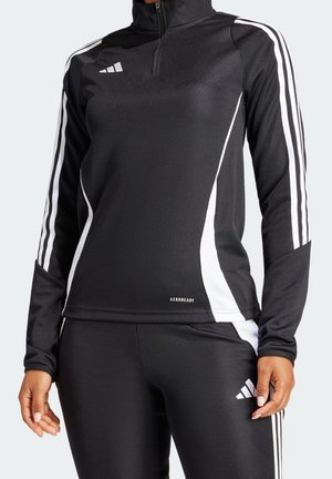 Femme portant un survêtement Adidas noir avec des bandes blanches sur les manches et les pantalons, col zippé et logo Aeroready sur le haut.