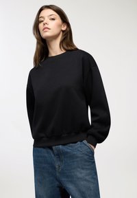 Giovane donna con capelli castani che indossa una felpa nera a girocollo e jeans blu, in piedi con una mano in tasca contro uno sfondo bianco.